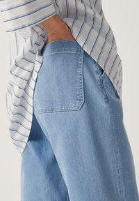 Světle modré džíny z denimu se střihem do rovna, s zadní kapsou a jemnou vybledlou texturou, kombinované s proužkovanou košilí viditelnou ze strany.