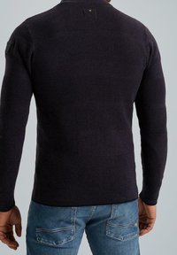 Pull en maille violet foncé avec une texture textile à côtes horizontales. Présente un col montant et un écusson avec logo sur l'épaule gauche.