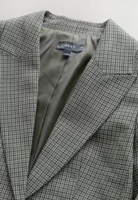 Hall ruuduline bleiser, millel on sile roheline vooder, tähtsa krae ja struktureeritud õlad. Silt kannab nime "NEXT".