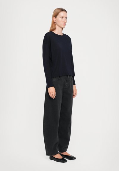 Tumedad sweater pikka varrukatega, sobitatud lõdva musta teksapüksiga. Mudel seisab kandmas lamedaid jalanõusid, minimalistlik disain, sile tekstuur, nähtavad mustrid puuduvad.