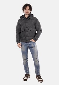 Giacca grigia intrecciata con chiusura a bottoni anteriori, due tasche sul petto e cappuccio; indossata sopra jeans blu sbiaditi e scarpe da ginnastica marroni.