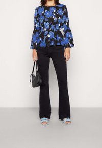 Black floral blouse with blue flowers, flared sleeves, and elastic waistband, paired with black flared jeans and light blue knotted sandals.