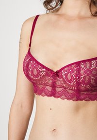 Spets-bh i djup rosa med blommönster, justerbara band, vågig nederkant och en liten rosett i mitten.