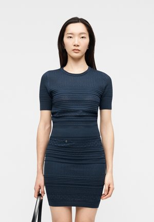 Giovane donna con lunghi capelli scuri che indossa un vestito in maglia blu navy aderente a maniche corte con trama, tenendo una borsa nera.