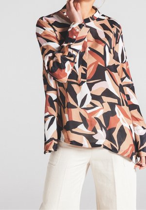 Femme portant un chemisier à manches longues avec des motifs abstraits marron, noir et blanc et un pantalon crème sur un fond clair uni.