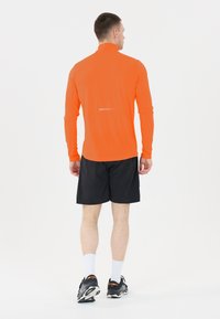 Oranssi pitkähihainen urheilupaita korkealla kauluksella, yhdistetty mustiin shortseihin. Heijastavat yksityiskohdat selässä, käytetään mustien lenkkarien ja valkoisten sukkien kanssa.