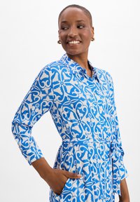 Blauwe jurk met patroon, voorzien van een kraag, lange mouwen en een riem op de taille. De stof is soepel met witte ingewikkelde ontwerpen en zakken.