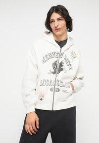Felpa bianca con zip, caratterizzata da una grafica "Heaven Sent Los Angeles", design di un'aquila e illustrazioni sulle maniche. Tessuto morbido, tasche frontali.