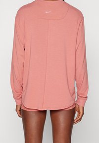 Langärmliges Sportoberteil in Korallenfarbe aus weichem Stoff, mit rundem Ausschnitt, angesetzten Schultern und kleinem weißem Logo auf der Rückseite.