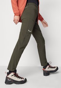 Calças de caminhada verde-oliva com um corte slim, apresentando bolsos com zíper e uma textura padrão. Combinadas com botas de caminhada pretas e cinzas.