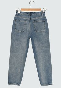 Jeans de mezclilla azul claro con bolsillos traseros colgando de una percha de madera contra un fondo liso.