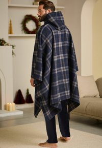 Marineblaues, kariertes Decken-Poncho mit Kapuze, das graue und braune Akzente aufweist. Weicher, strukturierter Stoff umschmeichelt marineblaue Hosen und lässt barfüßige Füße sichtbar.