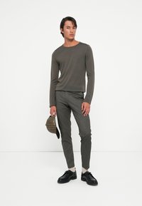 Long-sleeve olive green sweater, gray slim-fit trousers, black shoes, and a textured cap in plaid pattern held in one hand.