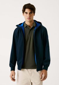 Mornarska zip-up jakna z modrim podloženjem kapuce, s poenostavljenim dizajnom in dolgimi rokavi. Obuta čez olivno polo majico.