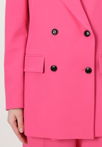 Double-breasted pink blazer in a smooth fabric. Features black buttons and two front pockets, with tailored lapels and structured shoulders.
