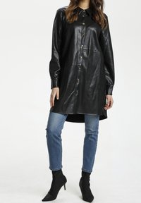 Abito camicia in pelle nera con chiusura a bottoni, maniche lunghe, fessure laterali, abbinato a jeans aderenti blu e stivaletti neri con tacco.