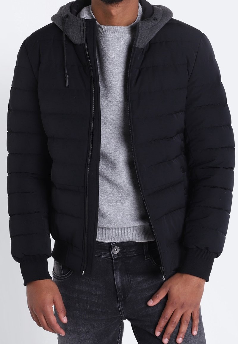 Zwarte gewatteerde jas met een grijze capuchon, ritssluiting, geribbelde manchetten en een eenvoudig ontwerp. Draagt over een grijze trui en donkere jeans.
