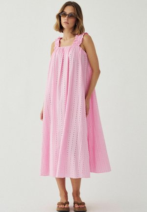 Žena v voľných, bezrúcnych ružových midi šatách s volánikovými ramienkami, textúrovaným materiálom, slnečnými okuliarmi a hnedými sandálmi, stojaca na svetlom pozadí.