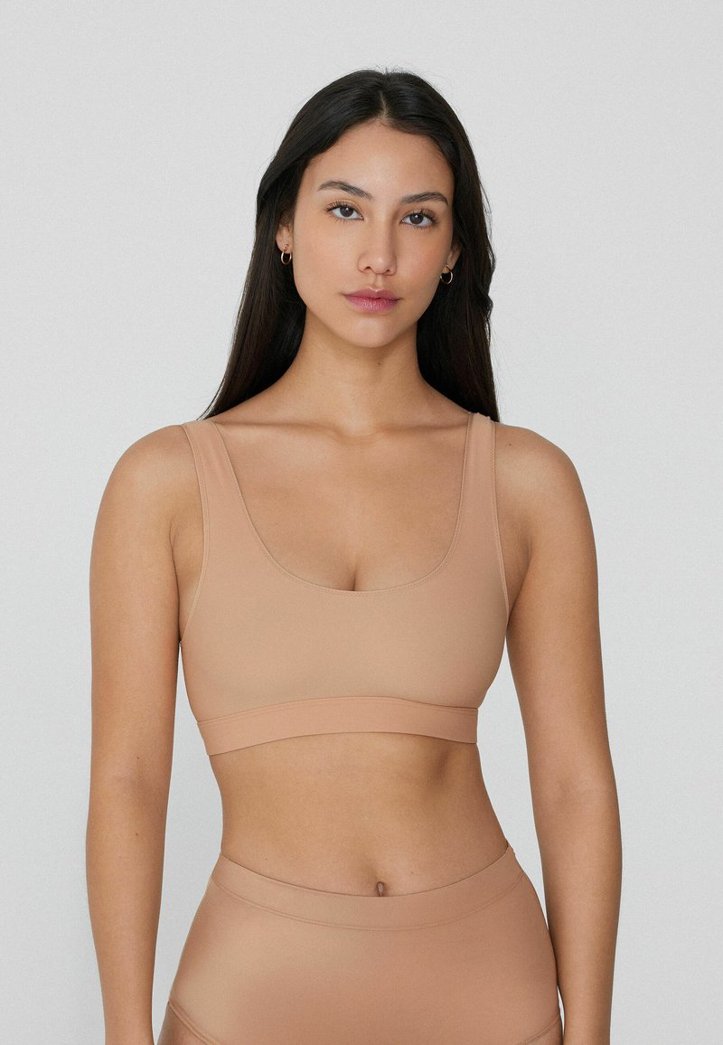 Hladká nude sportovní podprsenka s kulatým výstřihem a širokými ramínky. Měkký, elastický materiál s širokým pásem na spodní části pro podporu.