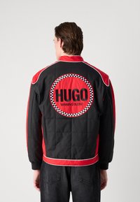 Czarna i czerwona pikowana kurtka bomberka z dużym, okrągłym logo "HUGO WINNING BLEND" na plecach, z kontrolowanymi akcentami i ściągaczami przy mankietach.