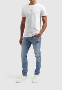 Witte katoenen t-shirt met "PURE PARTY" in het zwart gedrukt, gecombineerd met lichtblauwe denim jeans en witte sneakers.