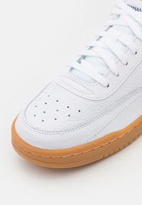 Bílé kožené tenisky s perforovanou špičkou, světle modrými tkaničkami a gumovou podrážkou. Mají minimalistický design a strukturovaný povrch.