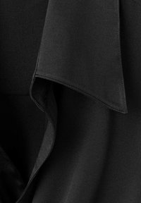 Čierna blúzka s hladkou textúrou, so splývavým golierom a čistými, preloženými okrajmi. Tkanina je ľahká a mierne lesklá.