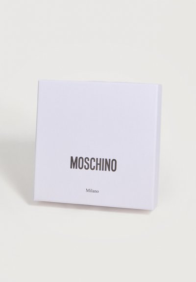Boîte carrée blanche avec "MOSCHINO" en lettres noires en gras et "Milano" en plus petit en dessous, sur un fond clair uni.