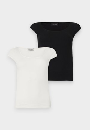 Vrouw staat en lacht, draagt een wit shirt met korte mouwen, grijze broek en zwarte loafers met hak, handen achter de rug, witte achtergrond.