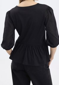 Haut noir avec des manches longues en tulle bouffantes et une taille peplum. Tissu lisse, encolure ronde et détail froncé à la taille.