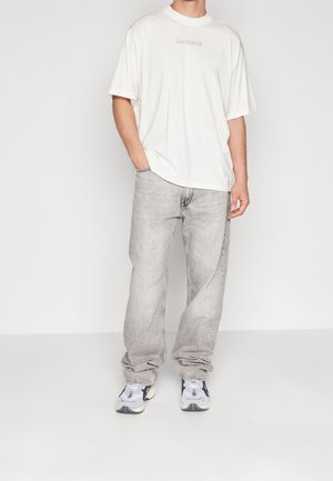 Hombre vistiendo una camiseta blanca de Lacoste, jeans sueltos de color gris claro y zapatillas blancas con detalles en negro y morado, de pie con una mano en el bolsillo.