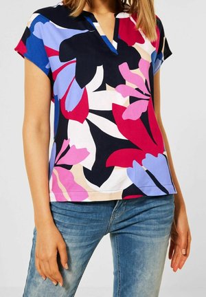 Kvinde iført en kortærmet V-hals bluse med store røde, pink, blå og sorte blomsterprint kombineret med blå jeans.