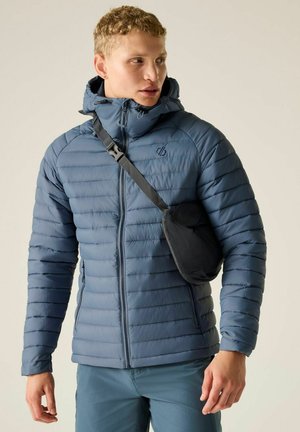 Young man wearing a zipped blue puffer jacket with a hood and a black crossbody bag, looking to the side against a plain background.