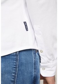 Camicia bianca a maniche lunghe in tessuto granuloso, con etichetta di marca sul lato e dettagli con bottoni sui polsini. Visibili jeans in denim blu.