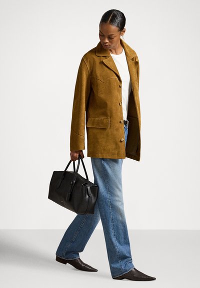 Nő barna velúr dzsekiben, fehér ingben, kék farmerben, fekete hegyes cipőben, fekete bőr kézitáskát tartva, lefelé fordított fejjel sétál.