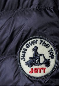 Il materiale della giacca puffa blu navy presenta una toppa circolare ricamata con il testo "Just Over The Top JOTT", che raffigura una figura su uno scooter.