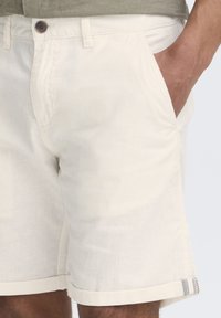 Pantalones cortos de color beige claro, elaborados con una mezcla de lino, con bolsillos laterales y un dobladillo vuelto con un acento a rayas. Cierre clásico con botón.