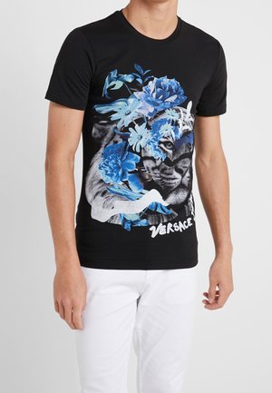 Homme portant un t-shirt noir avec un motif coloré de fleurs bleues et un visage de tigre, accompagné du logo blanc "Versace", associé à un pantalon blanc.