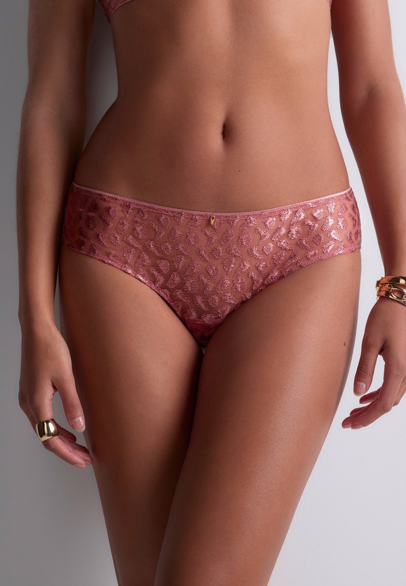 Femme portant des sous-vêtements en dentelle semi-transparente avec imprimé léopard rose, une bague en or à la main gauche et des bracelets en or au poignet droit, se tenant devant un fond gris.