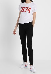 Camiseta blanca con estampado rojo "1974", combinada con jeans skinny negros y zapatillas blancas con acentos rojos y azules.