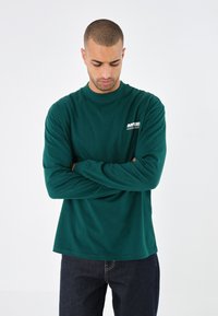 Grünes Langarmshirt aus weichem Stoff mit rundem Halsausschnitt und weißem Logo-Detail auf der Brust, kombiniert mit dunkler Jeans.
