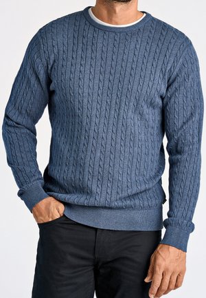 Hombre con suéter azul de punto trenzado sobre una camiseta blanca y pantalones negros, con una mano en el bolsillo y la otra relajada.