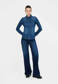 Dunkelblaues Jeanshemd mit tailliertem Schnitt, zwei Brusttaschen und Knopfleiste. Kombiniert mit dunkelblauen Jeanshosen mit weitem Bein.