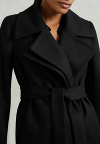 Naine kannab musta vöödiga villakatet, millel on laiad kraed ja sügav V-kujuline kaelus, neutraalses hallis taustal.