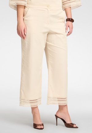 Beige Hosen mit weitem Bein und Spitzenbesatz am Saum, kombiniert mit braunen, hochhackigen, offenen Mules und einem passenden Armband am rechten Handgelenk.