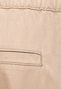 Μπεζ ύφασμα με ελαστική μέση και μία οριζόντια τσέπη με σχισμή και εμφανείς ραφές.