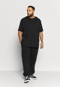 Chemise noire à manches courtes avec des rayures verticales blanches, associée à un pantalon noir et des baskets noires. Design simple et ample, texture de tissu lisse.