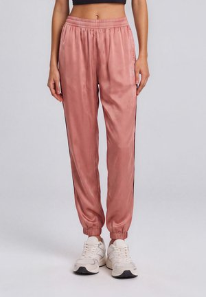 Femme portant un pantalon de jogging en satin rose avec taille et poignets élastiques, associé à des baskets blanches, debout sur un fond uni.