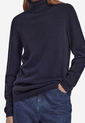 Femme portant un pull à col roulé bleu marine foncé à manches longues et un jean en denim bleu, une main dans la poche de manière décontractée.