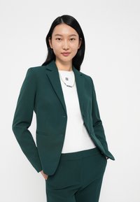 Tmavě zelený přizpůsobený blejzr s klopami, dvěma předními kapsami a slim fit střihem, nošený přes bílý top s brandeingem "HUGO".